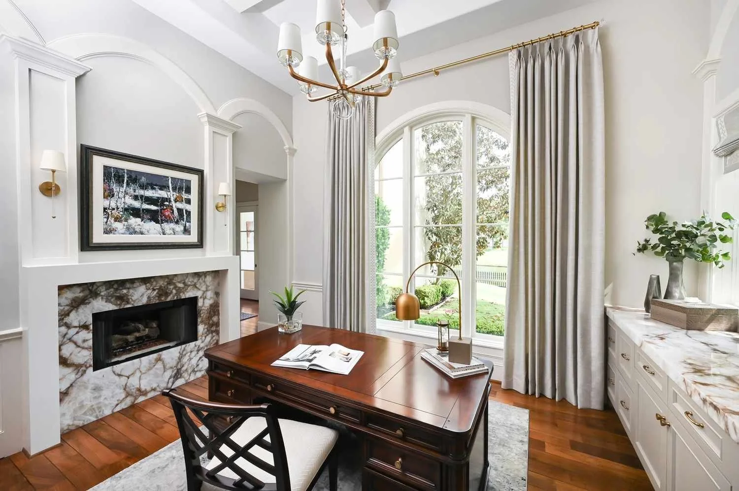 Bright and airy office with a wooden desk and fireplace, featuring natural lighting from a big window.